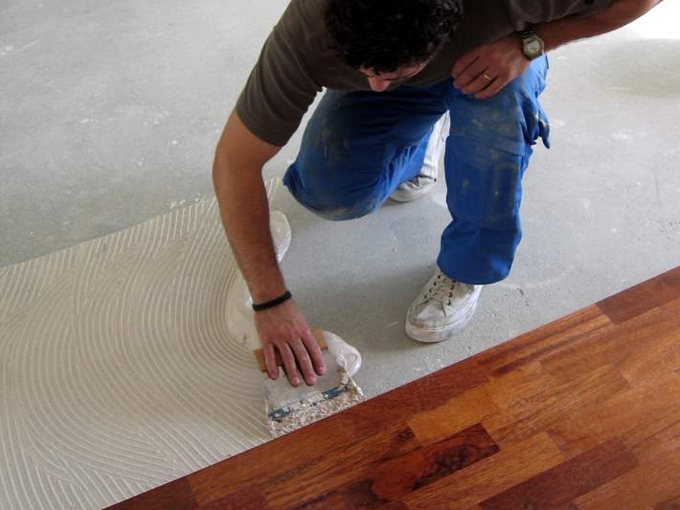 Installer spreading adhesive on a concrete slab with a notched trowel while laying timber parquet